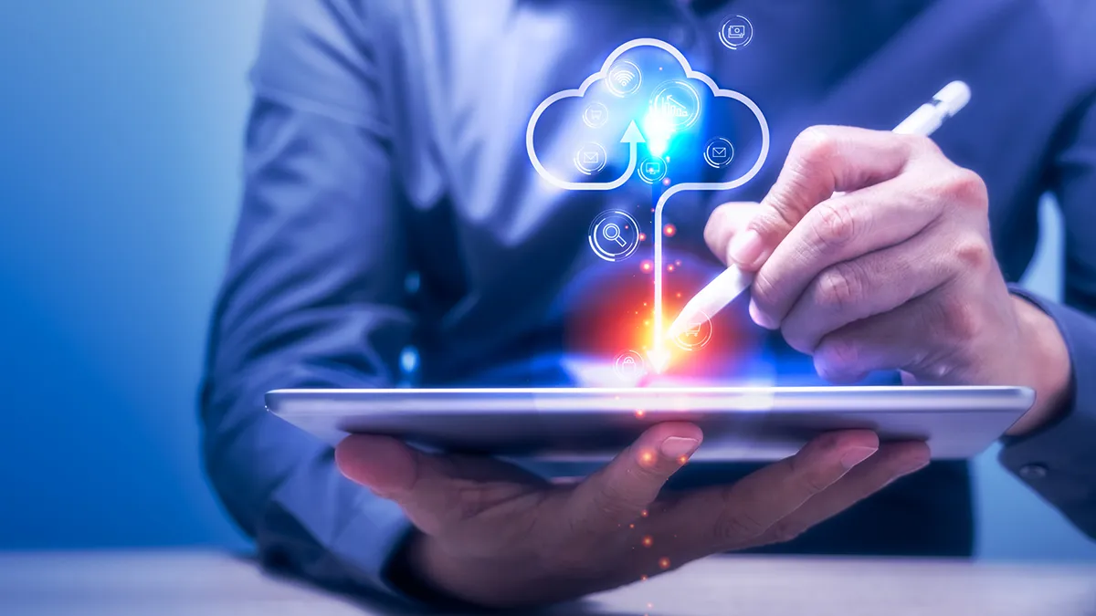 A person in a business suit holding a tablet with a stylus. The tablet screen displays a cloud graphic surrounded by various icons, including currency symbols and upward-pointing arrows, suggesting growth. The background is blurred with a blue tint, focusing attention on the hands, tablet, and the graphical representation of cloud services. Image for a blog article: How to achieve AWS cloud cost optimization with FinOps?