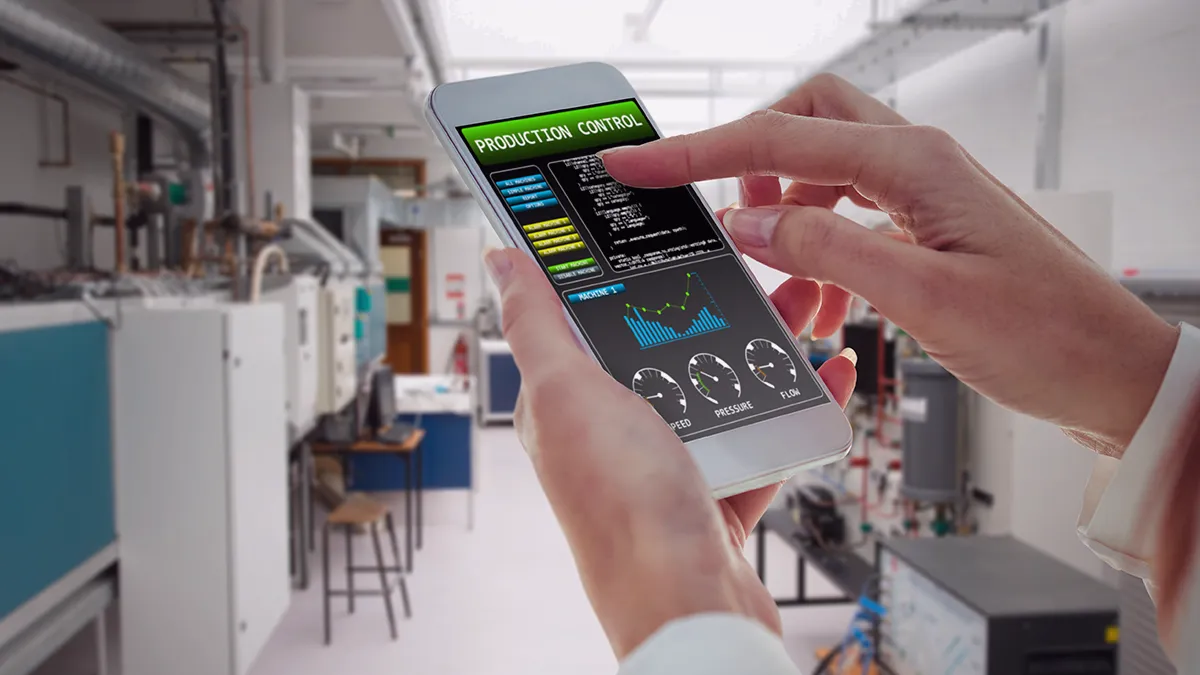 The photo shows a woman's well-groomed hands holding a smartphone, and in the background we see a technical room with specialized equipment. The phone's monitor shows the words “Production Control” and graphs. From the context, it appears that the woman is checking the parameters of the production line. The photo is the cover graphic for an expert article on Transition Technologies PSC's blog, and appears as an illustration of a post titled: What is Predictive Maintenance in Industry 4.0? – solution for Smart Factory.