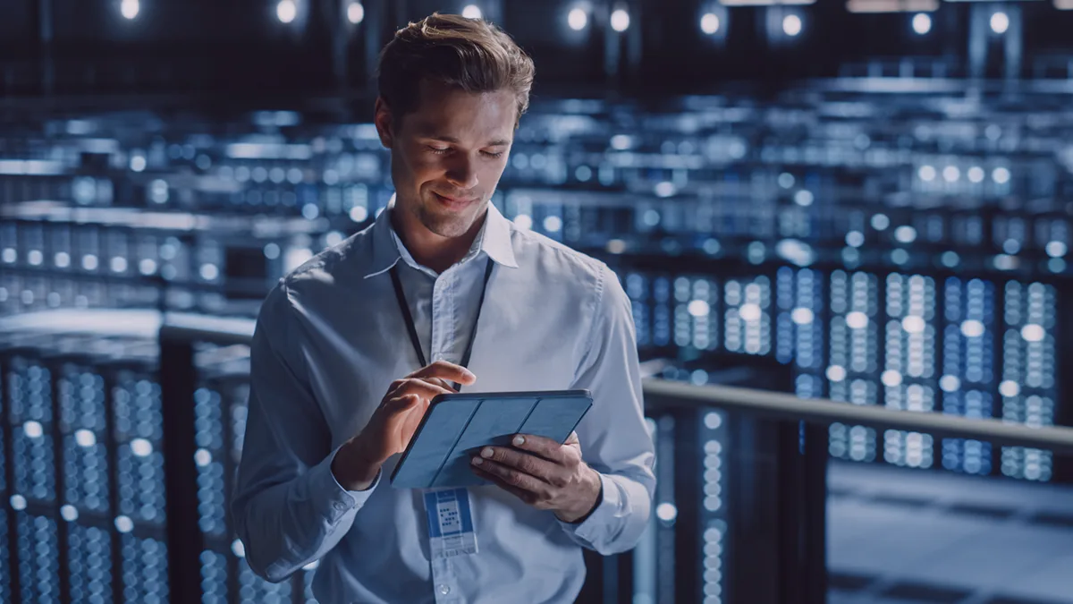 The photo shows a young man standing in the background of a server room; he is holding a tablet in his hand, at which he is directing his gaze, with his hand hovering over the tablet's screen; the man is dressed in a long-sleeved shirt, and also has a company badge around his neck. The photo is the cover image for a Transition Technologies PSC blog post titled: Digital Maintenance: Two birds with one stone