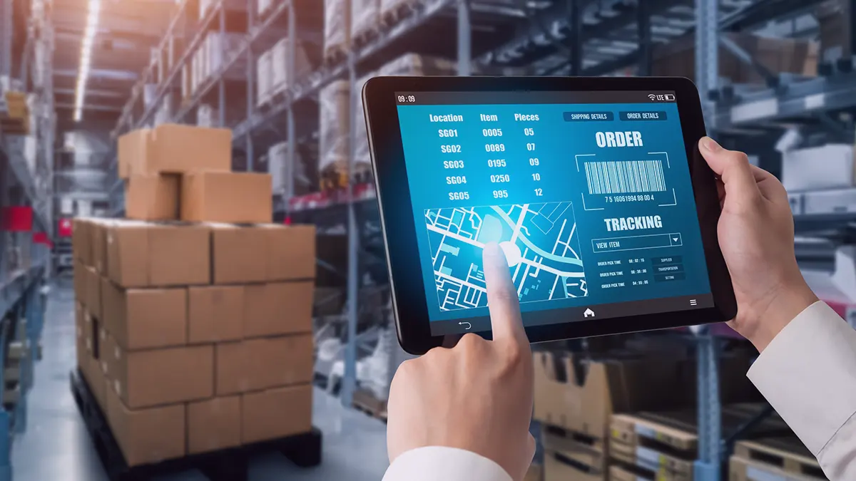 The photo, which is the cover of the Transition Technologies PSC blog post, shows a tablet held in the hands visible in the lower right corner, the rest of the silhouette is invisible; the device's screen displays order data, counts, a map and a QR code, in the background you can see a pallet of cardboard boxes, from the context it seems that these are goods packed for shipping; in the background you can see metal warehouse shelves, full of various products.