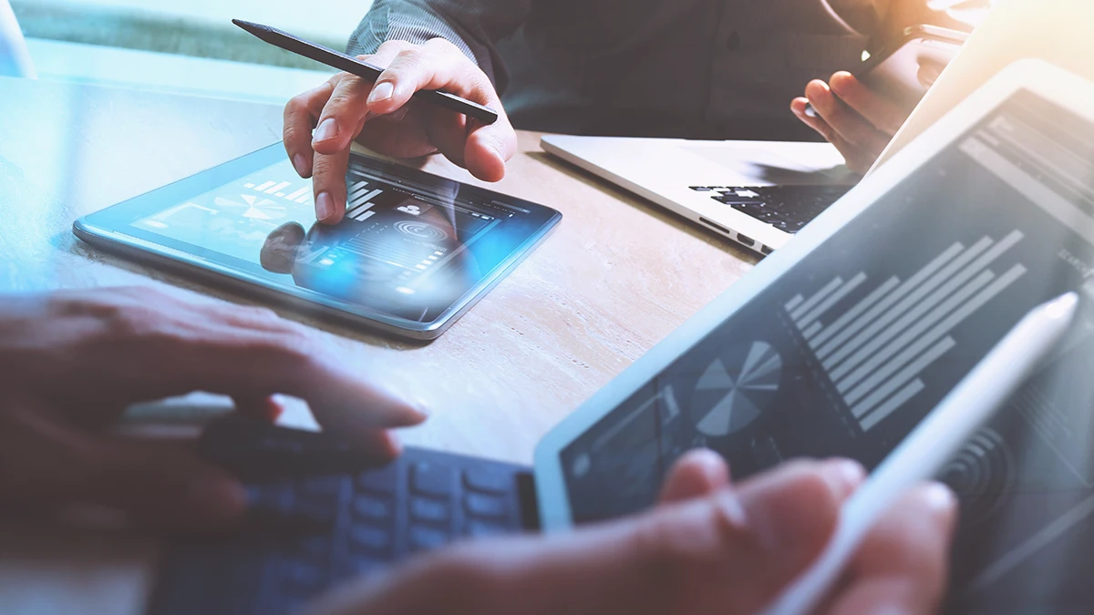 Zdjęcie przedstawiające blat biurka i dwa tablety z uniesionymi nad nimi dłońmi; na ekranach są widoczne wykresy, z kontekstu zdjęcia wynika, że jest to spotkanie w trakcie pracy w celu omówienia wyników. Zdjęcie stanowi okładkę wpisu blogowego firmy Transition Technologies PSC o tytule: Optymalizuj zapasy i oszczędzaj dzięki trafnemu prognozowaniu popytu w handlu detalicznym