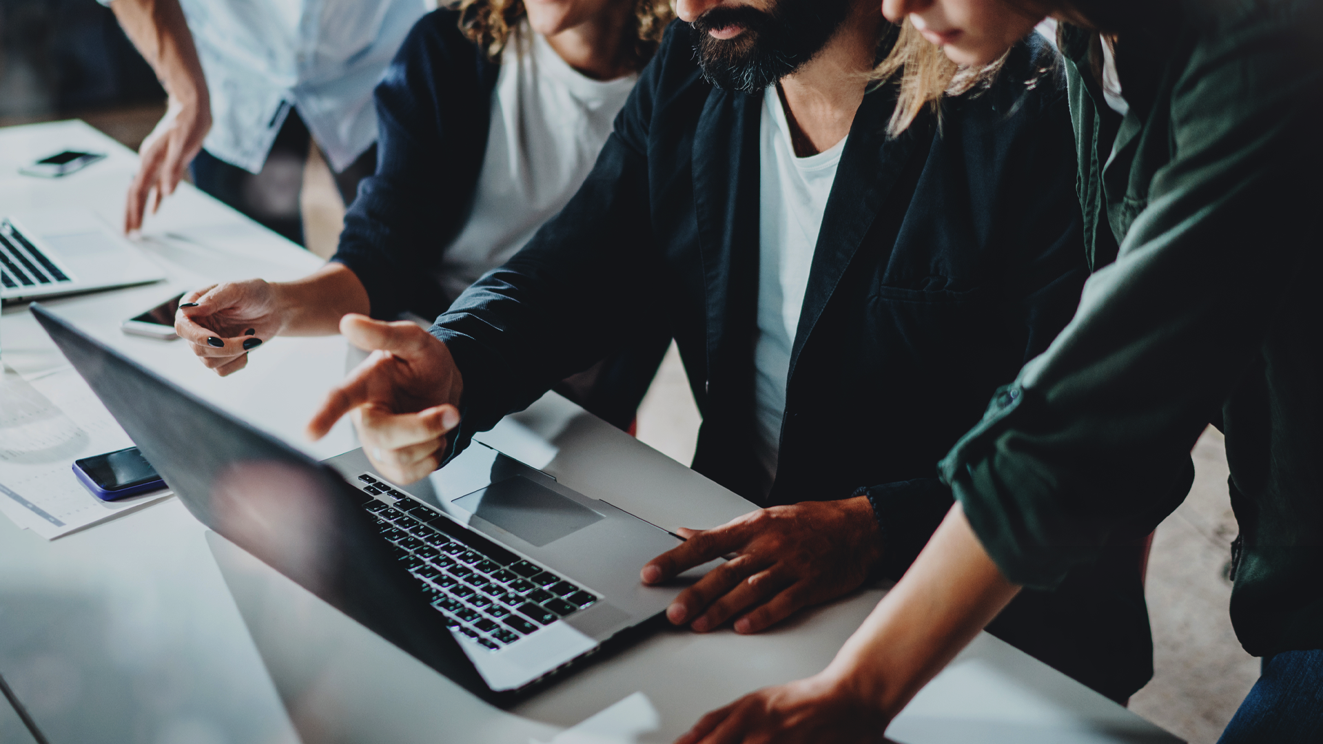 Zdjęcie przedstawiające grupę ludzi pochyloną nad otwartym laptopem, wskazujących na jego ekran; na zdjęciu widoczne są tylko biurko, tułowia i ręce postaci; otoczenie jest biurowe, z kontekstu wynika, że zespół omawia wspólnie jakąś nagłą usterkę systemową. Zdjęcie jest fotografią okładkową do artykułu blogowego firmy Transition Technologies PSC