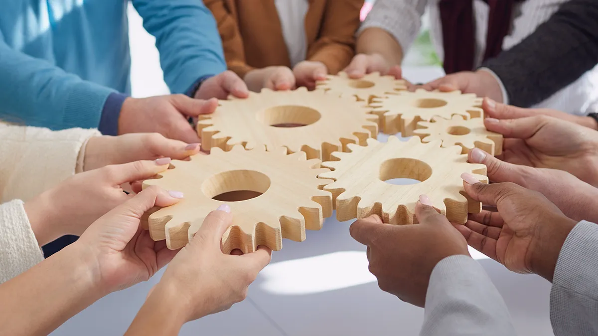 Zdjęcie przedstawia grupę ludzi ustawionych w okręgu, a właściwie ich dłonie trzymające drewniane koła zębate. Koła zazębiają się ze sobą przyłożone do siebie po środku kadru. Z kontaktu wynika, że zdjęcie symbolizuje udaną współpracę, dzięki której każdy element jest na swoim miejscu i wszystko działa prawidłowo. Zdjęcie jest grafiką okładkową do notki prasowej firmy Transition Technologies PSC i pojawia się jako ilustracja wpisu o tytule: Transition Technologies USA dołącza do Transition Technologies PSC.