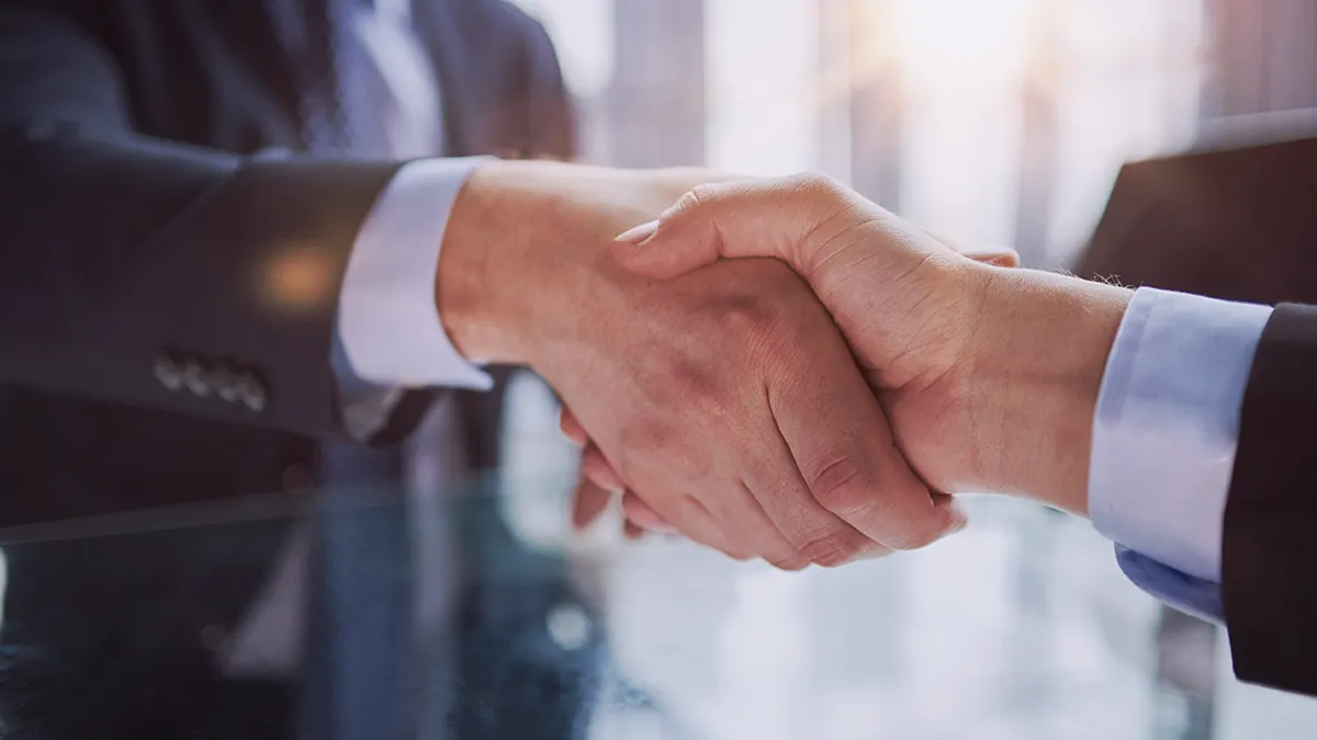 Une image en gros plan de deux personnes en tenue professionnelle se serrant la main, symbolisant un accord formel. La poignée de main est bien centrée, avec un arrière-plan de bureau flou éclairé par la lumière naturelle, indiquant un cadre professionnel. Cette image représente l'annonce de l'entrée en partenariat de Transition Technologies PSC avec GitHub, soulignant le début d'une relation de collaboration.