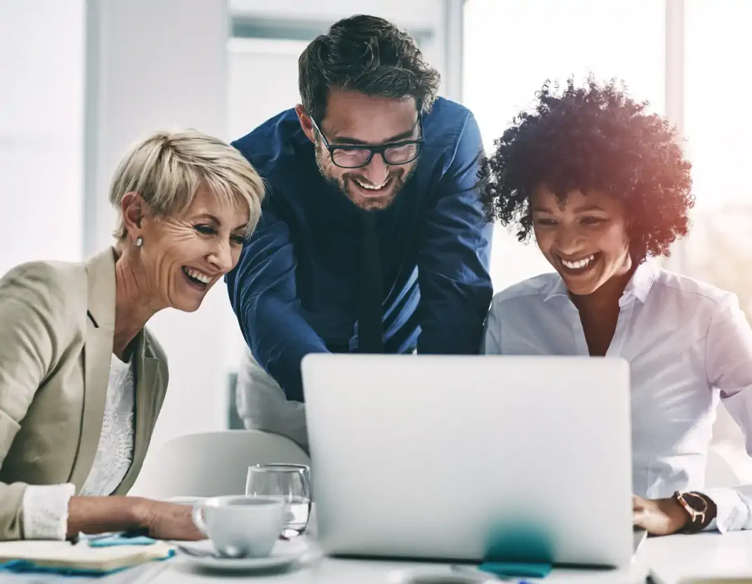 Three diverse business professionals smiling and collaborating around a laptop in a bright, modern office