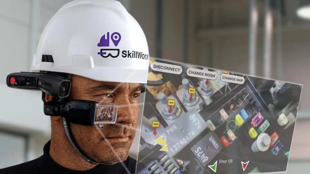 Zdjęcie przedstawia pracownika linii produkcyjnej, który ma na sobie kask ochronny z nowoczesnym, małym ekranem zamocowanym do kasku; ekran znajduje się przed prawym okiem pracownika i wyświetla obraz widoczny w kamerze urządzenia z naniesionymi oznaczeniami urządzeń. Dzięki temu pracownik wyświetla pomocniczą instrukcję bez użycia rąk. Komendy wydaje głosowo. Na kasku widnieje logo oprogramowania sprzętu - SkillWorx autorstwa firmy Transition Technologies PSC