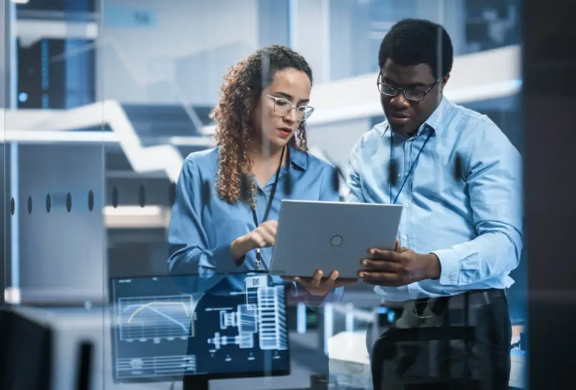 Two people discussing data on a laptop in a modern office, surrounded by screens displaying charts.