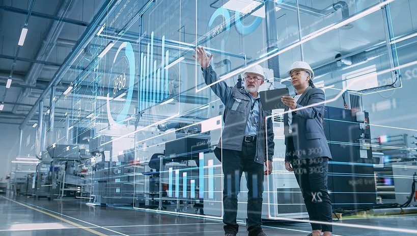 Zwei Industriefachleute in Schutzausrüstung stehen in einer Hightech-Fertigungsanlage und analysieren eine transparente digitale Schnittstelle, die Echtzeitdaten, Diagramme und Leistungsmetriken anzeigt. Die futuristische Überlagerung stellt intelligente Fabrikabläufe, digitale Zwillinge und datengesteuerte Entscheidungsfindung in industriellen IoT-Umgebungen